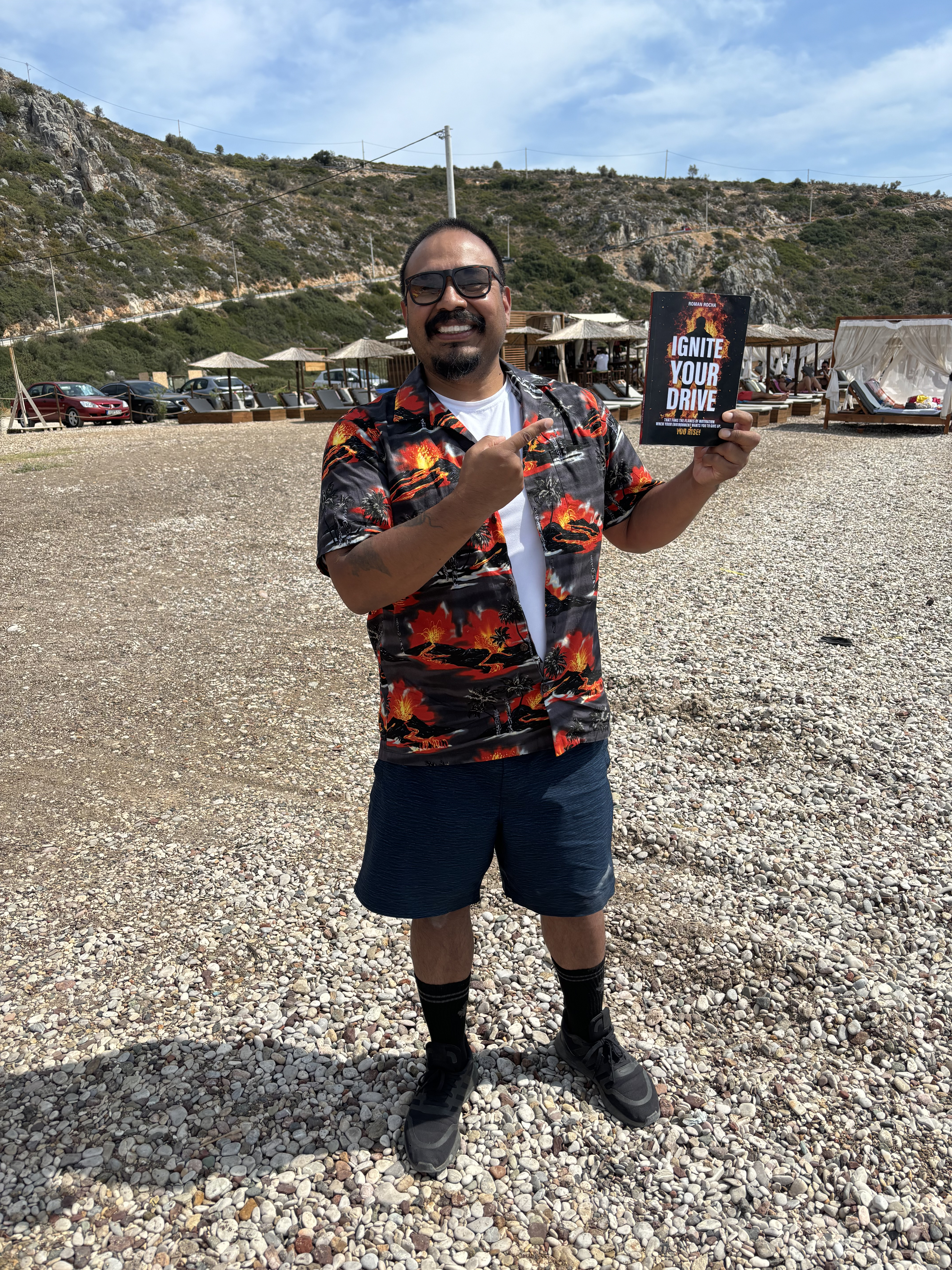 Roman Rocha holding his book titled Ignite Your Drive on the Beach in Greece